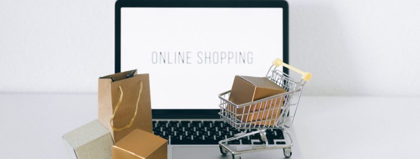 En laptop med texten "online shopping" på skärmen, små paket, en liten kundvagn och kasse står på tangentbordet